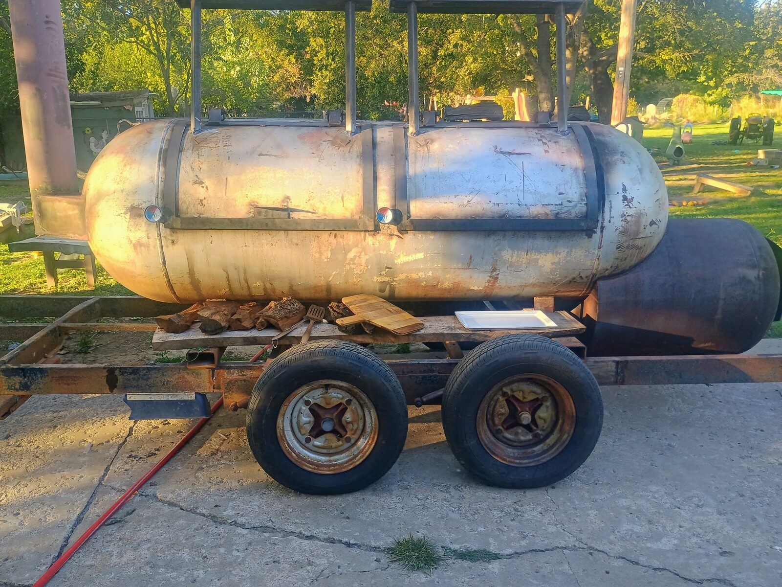 Smoker mounted on the trailer — unpainted, firebox and chimney visible, wood stacked on the shelf